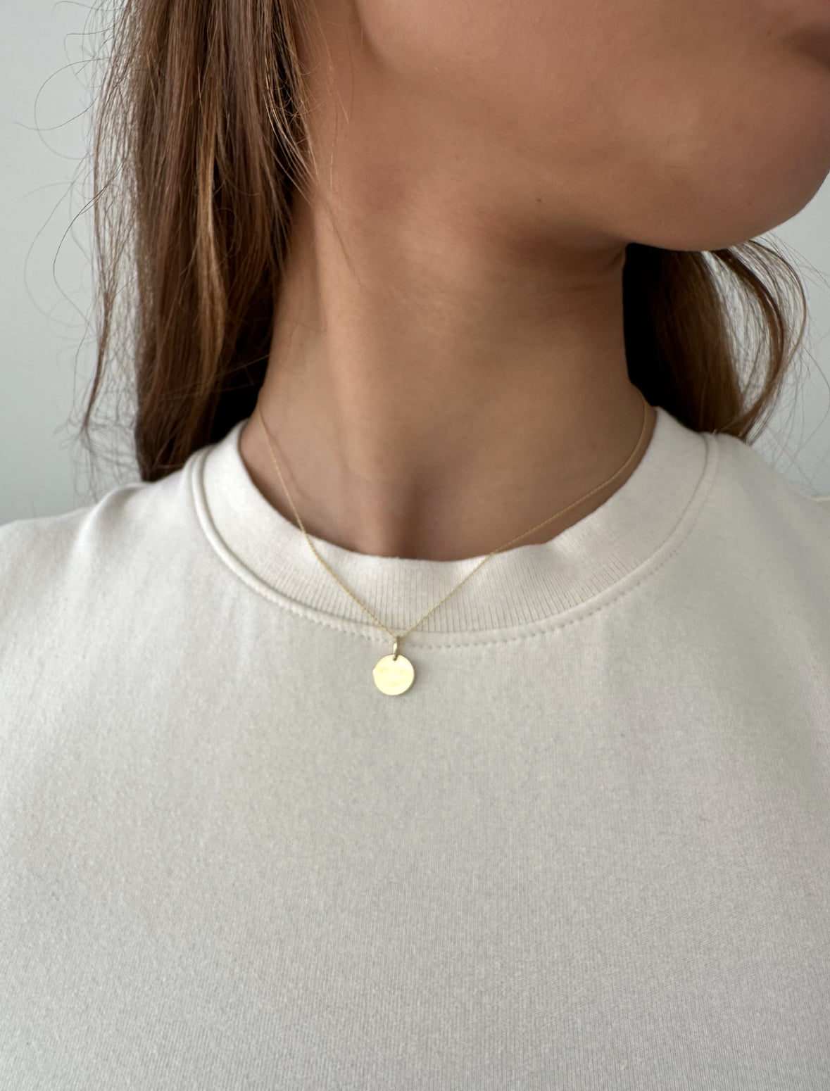 Ronde 14 karaat gouden hanger aan een gouden ketting gedragen door jonge vrouw met bruin haar in witte top, close-up