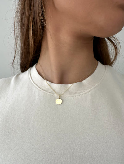 Ronde 14 karaat gouden hanger aan een gouden ketting gedragen door jonge vrouw met bruin haar in witte top, close-up